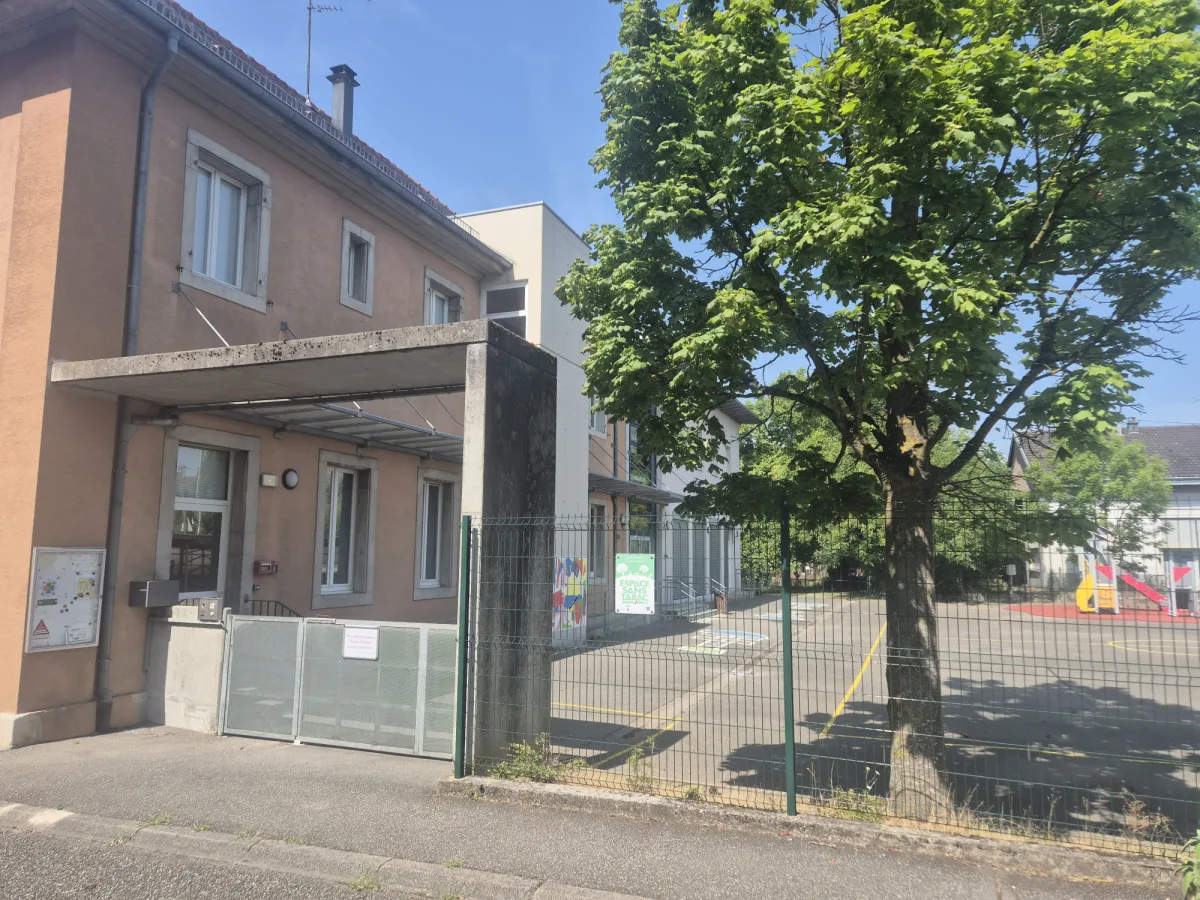 Façade de l'école Saint-Bernard vue depuis la rue, avec son préau en béton, son portail vitré et la cour de récréation clôturée.