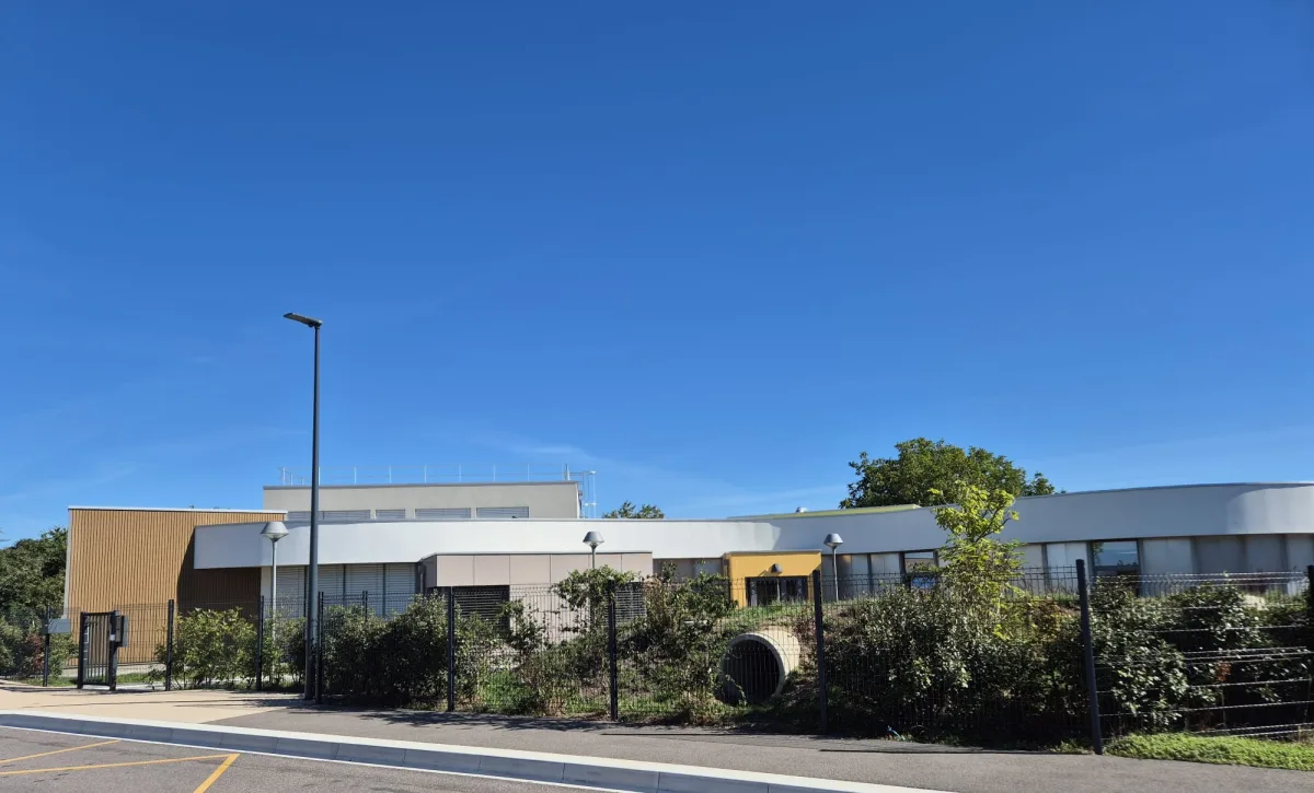 Le bâtiment périscolaire vu depuis la rue : bardage bois clair, façade blanche aux lignes courbes et entrée jaune, derrière une clôture.