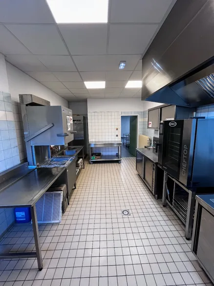 Cuisine professionnelle vue dans l'axe central, tables de travail en inox de part et d'autre, four mixte à chariot et hotte inox.