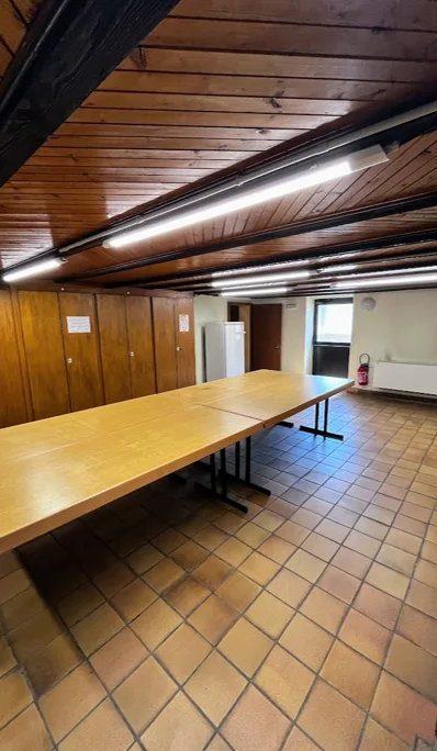 Caveau de la salle des fêtes avec plafond lambrissé, poutres apparentes, longues tables rectangulaires assemblées et sol carrelé de tomettes.
