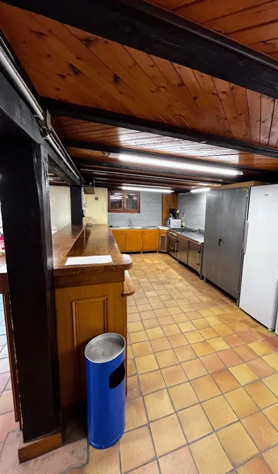 Cuisine de la salle des fêtes vue depuis le comptoir de bar en bois, avec plan de travail inox, meubles bas et grands réfrigérateurs.