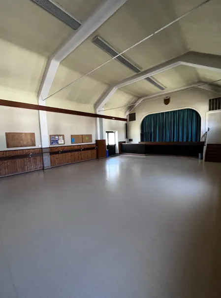 Grande salle des fêtes vide avec sol en résine gris clair, scène fermée par un rideau bleu, lambris muraux en bois et charpente blanche apparente.