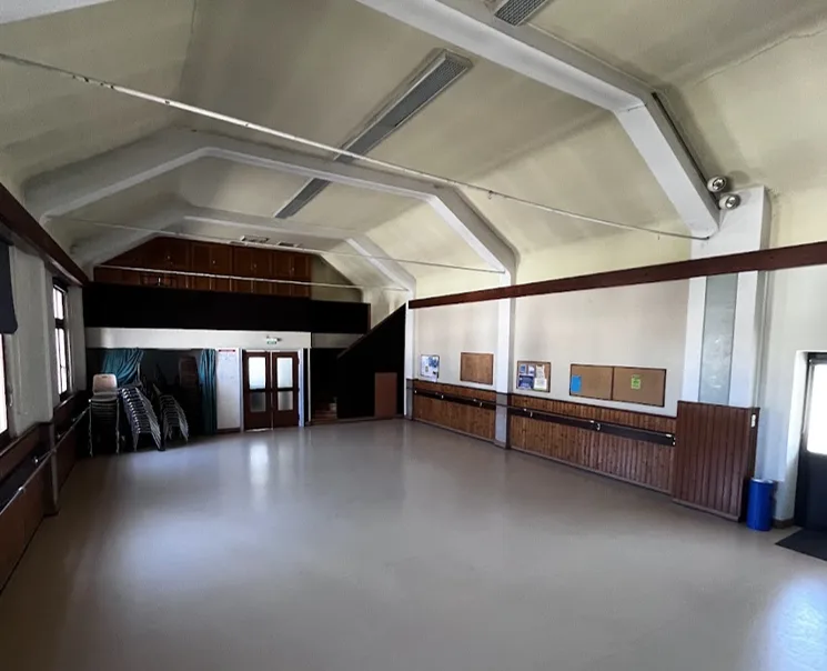 Salle des fêtes vue vers l'entrée, avec mezzanine en bois, portes vitrées à double battant et chaises empilées le long des lambris.
