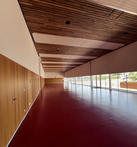 Salle rouge tout en longueur, sol résine bordeaux, plafond à lames de bois foncé, murs habillés de panneaux en bois et grande façade vitrée.