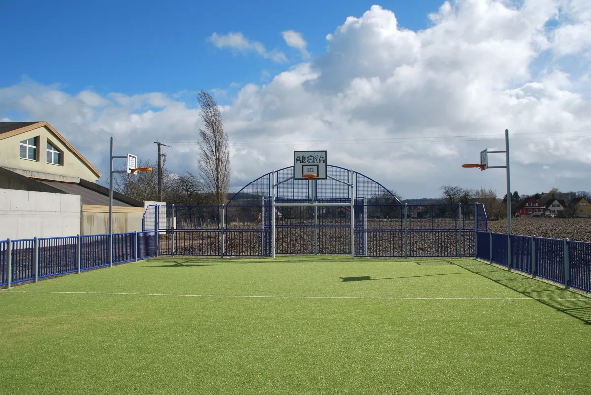 Le city-stade communal en gazon synthétique, entouré d'une clôture bleue, équipé de deux paniers de basket et de buts, en bordure de champs